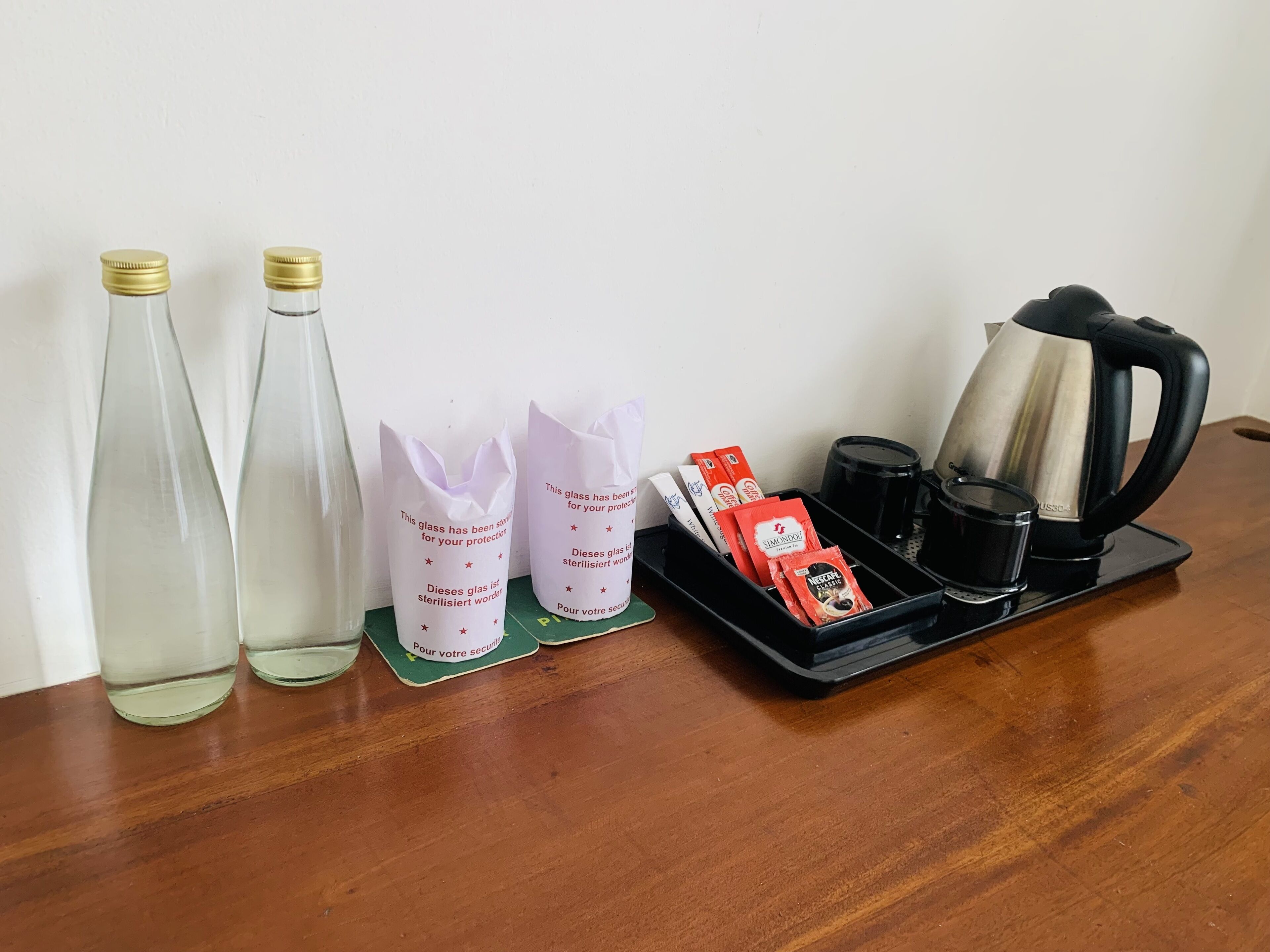 Dos botellas de vidrio, dos bolsas de papel con texto, una bandeja con paquetes de café, una cafetera y una taza sobre una mesa de madera.