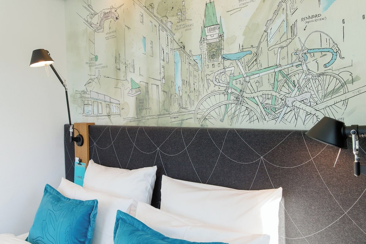 Habitación de hotel con cama, dos almohadas, cabecero con diseño geométrico, mapa mural y dos lámparas.