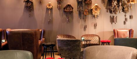 A room with multiple antique cuckoo clocks on the wall, leather armchairs, and a table with red spherical cushions.