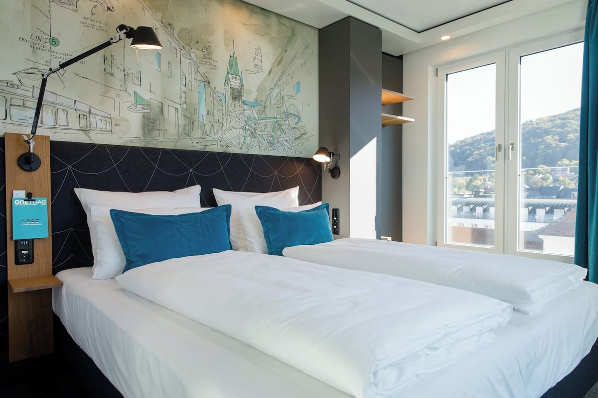 Habitación de hotel con una cama grande, cabecera con un mural de la ciudad, mesita de noche con lámpara, y vistas a un puente y colinas a través de la ventana.