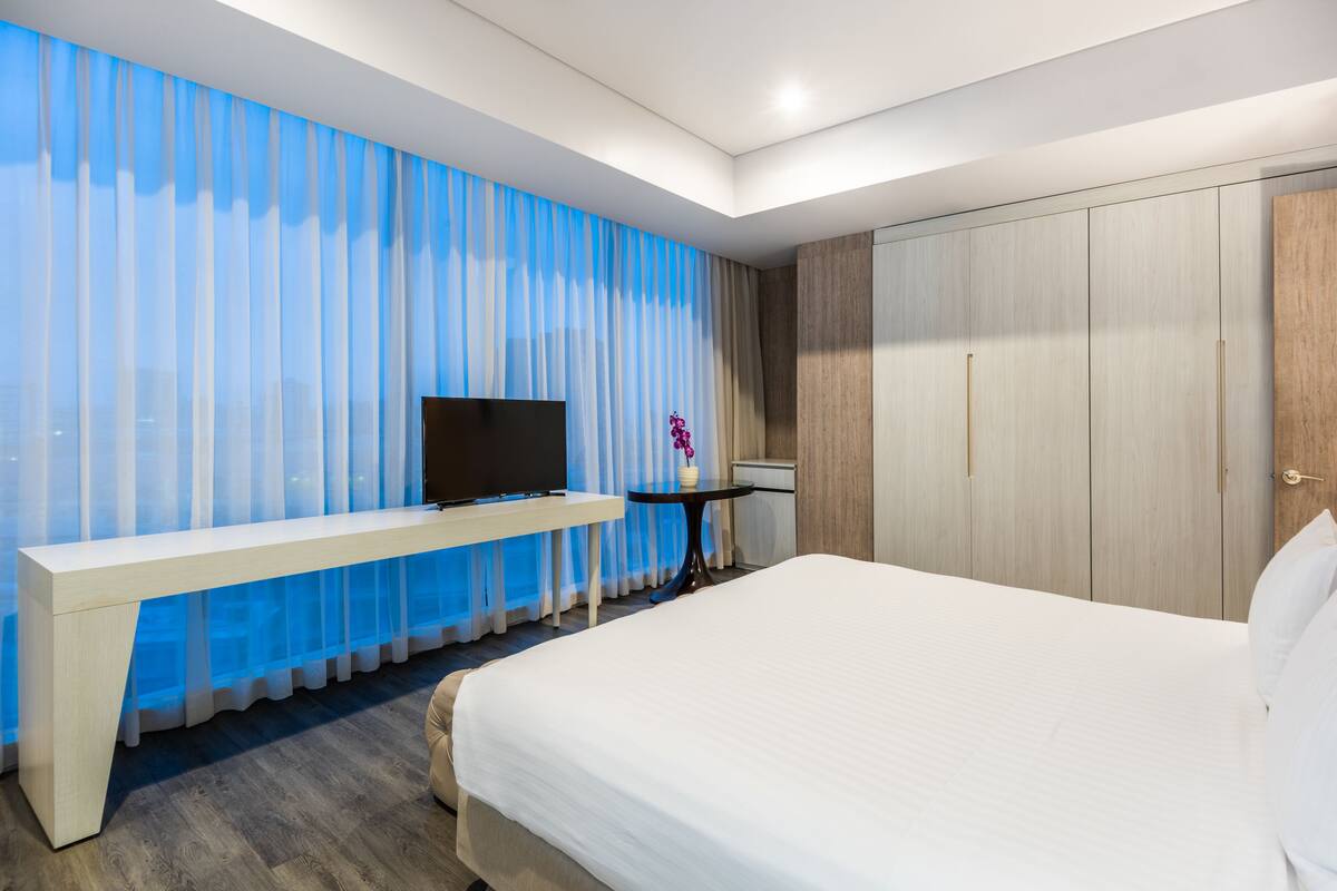 Une chambre d’hôtel avec un lit, un téléviseur posé sur une console blanche, une petite table ronde avec un vase et une armoire.