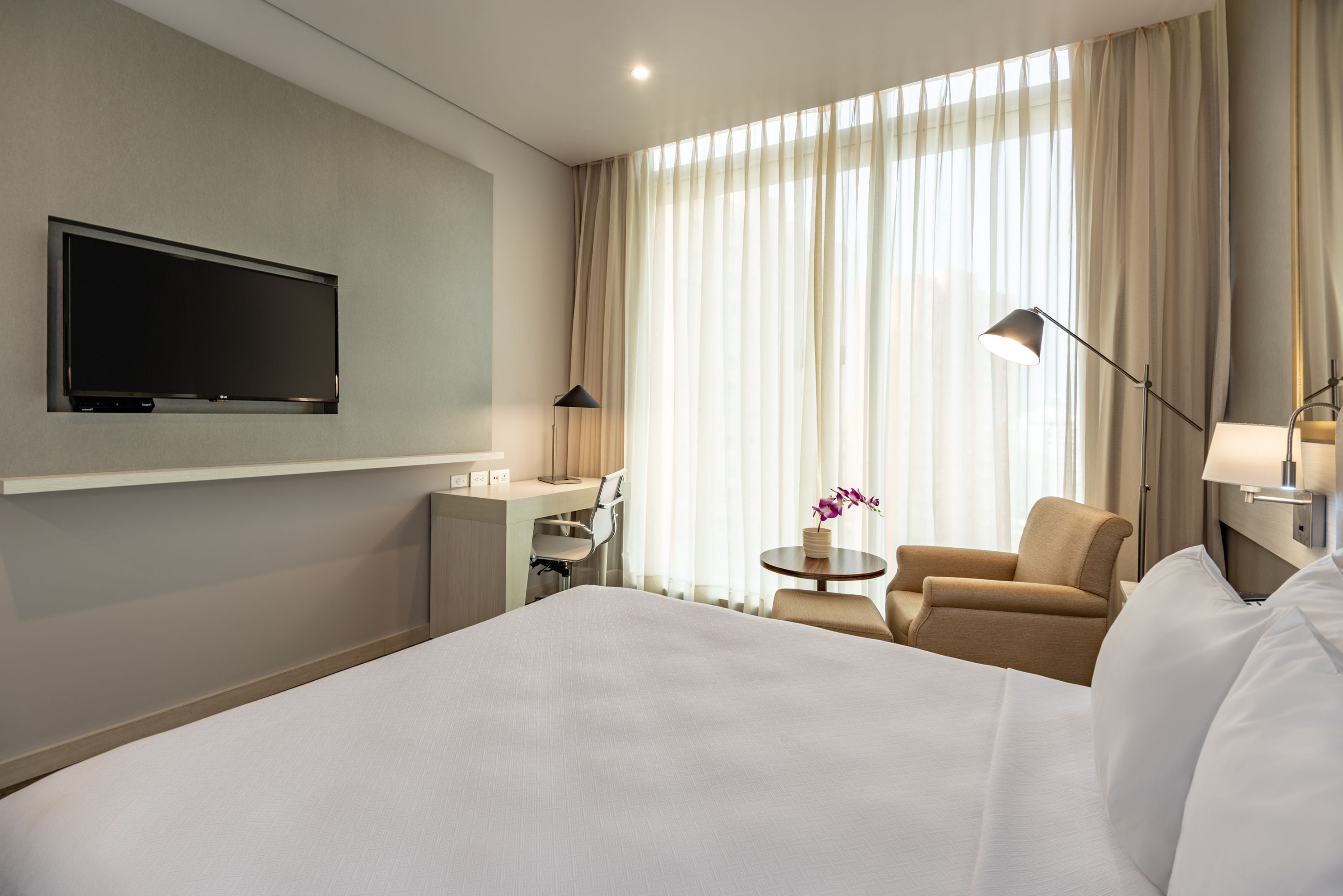 A hotel room with a large bed, a desk with a chair, a TV mounted on the wall, a lamp, and a window with curtains.