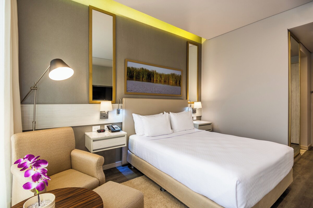 Een hotelkamer met een bed, nachtkastjes, een lamp, een stoel, een klein tafeltje met een orchidee en een schilderij aan de muur.
