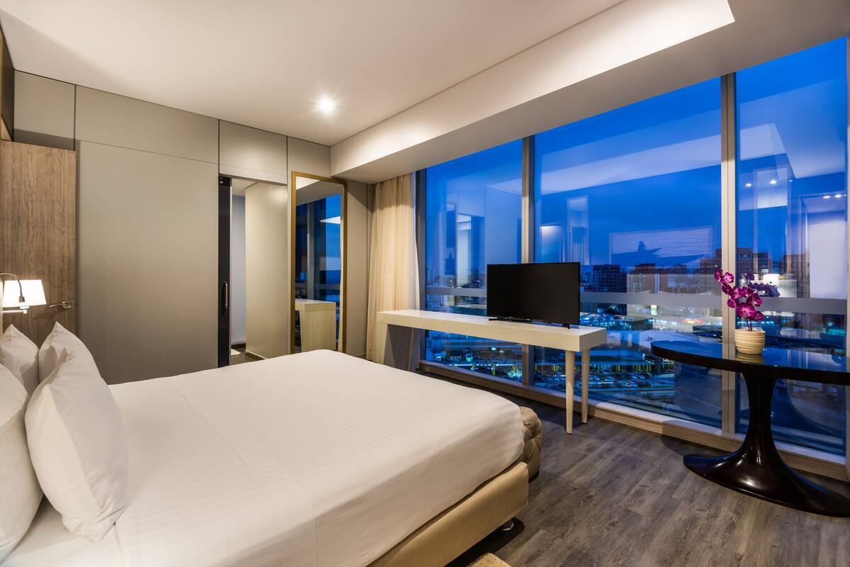 Une chambre d’hôtel moderne dotée d’un grand lit, d’un bureau avec une télévision et offrant une vue sur la ville grâce à de grandes fenêtres.