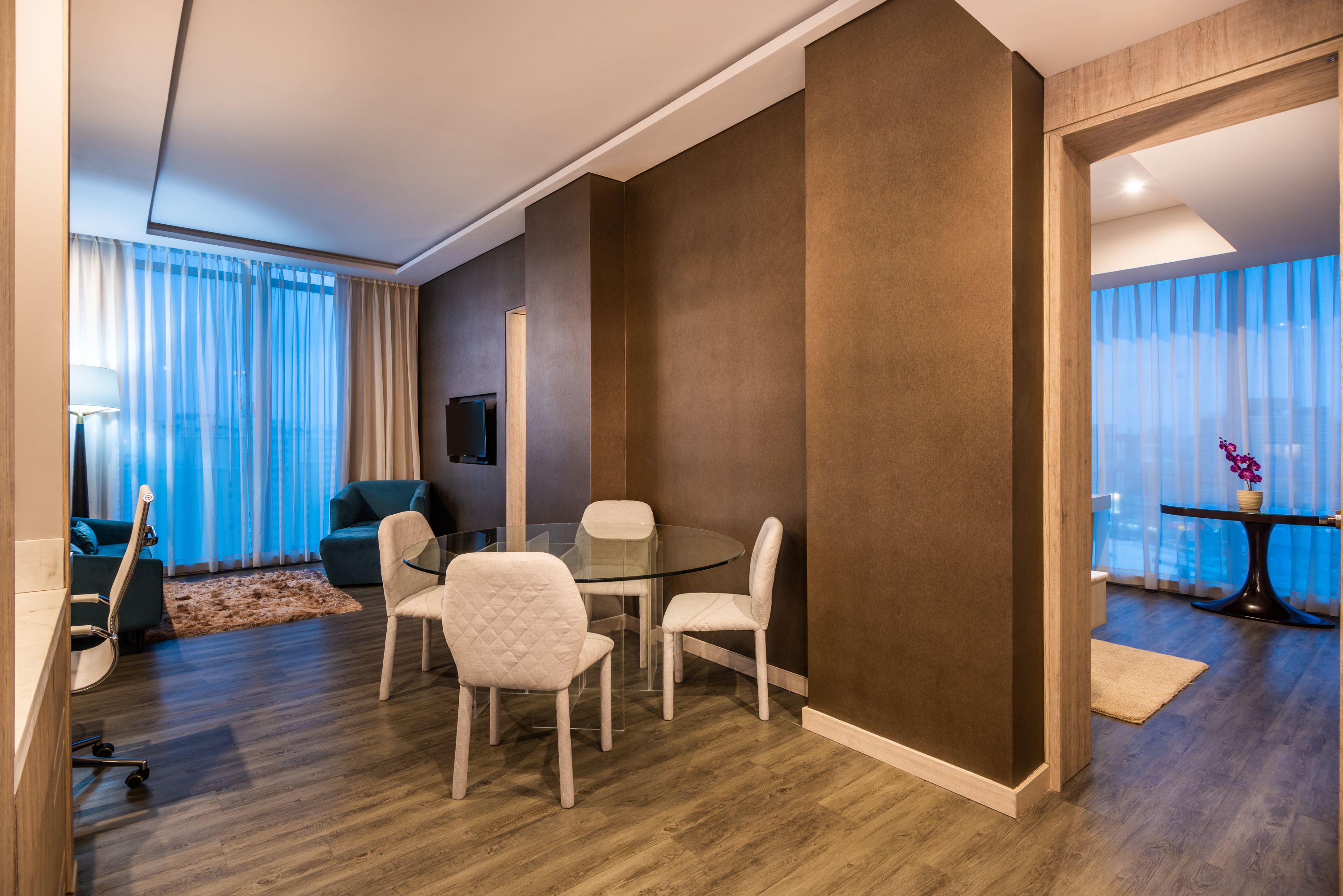A modern hotel room with a glass dining table, two chairs, a sofa, and a TV.