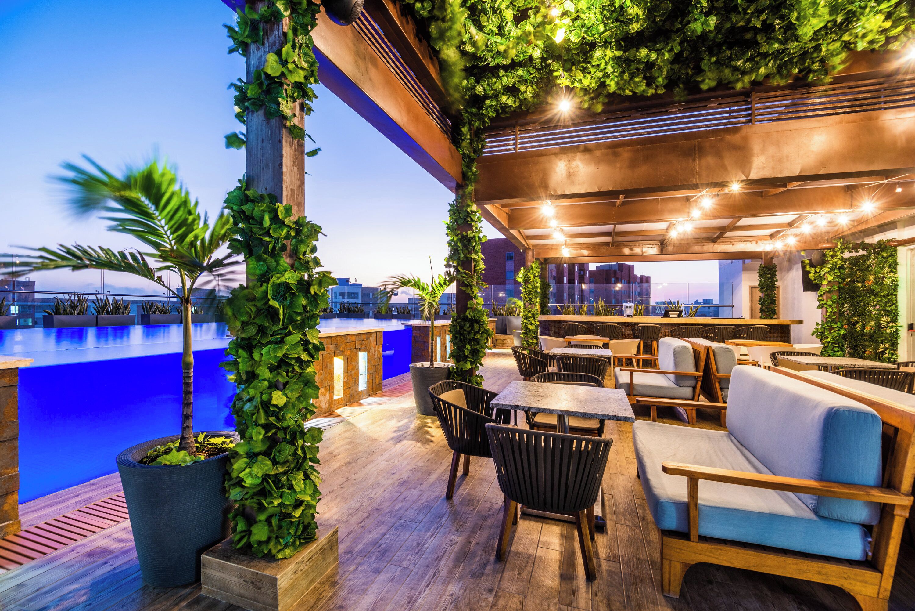 Un área de estar al aire libre con mesas y sillas de madera, rodeada de vegetación y vista a una piscina.