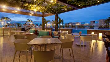 An outdoor seating area with tables and chairs, a pool, and a view of the city skyline at dusk.