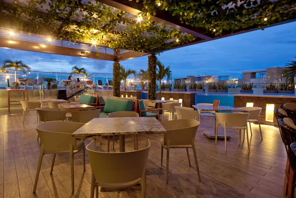 Un área de estar al aire libre con mesas y sillas, una piscina y vistas del horizonte urbano al anochecer.