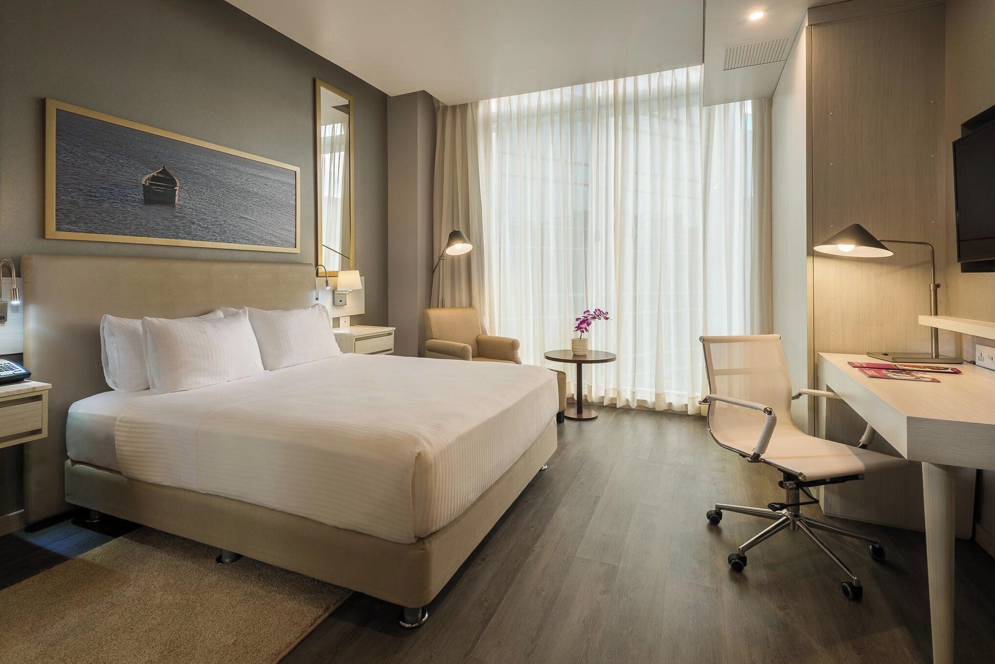 Habitación de hotel con una cama grande, un escritorio, una silla, una mesita con un jarrón de flores y un cuadro en la pared.