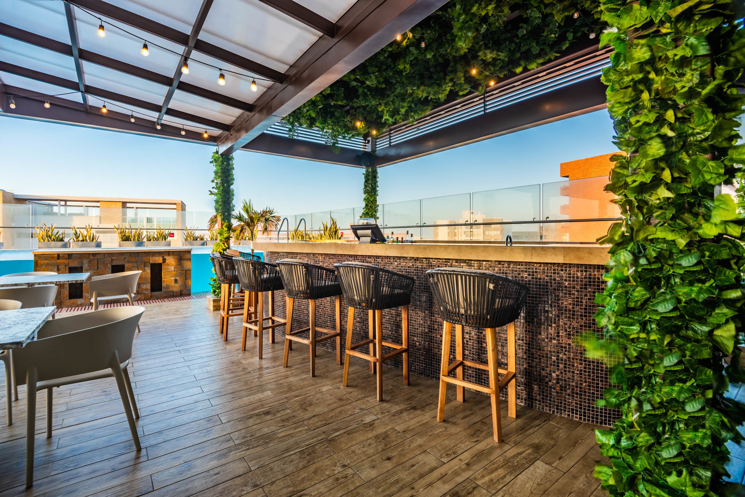Zona de bar al aire libre con suelo de madera, taburetes y barandilla de cristal.