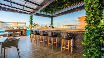 Zona de bar al aire libre con suelo de madera, taburetes y barandilla de cristal.