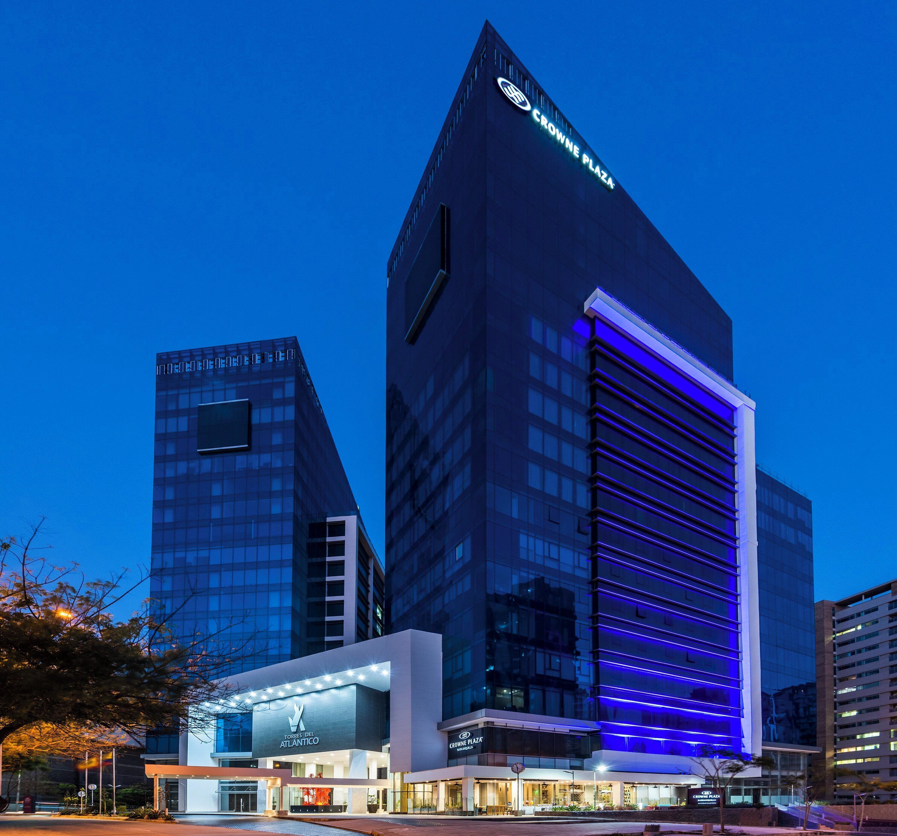 A modern hotel with a prominent 'Crowne Plaza' sign, featuring a sleek glass facade and a distinctive blue-lit entrance.