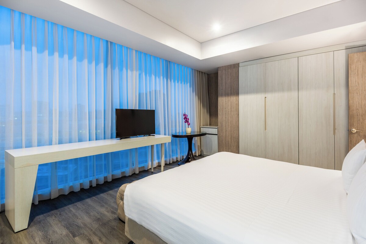 Een hotelkamer met een bed, een televisie op een witte console, een klein rond tafeltje met een vaas, en een kledingkast.