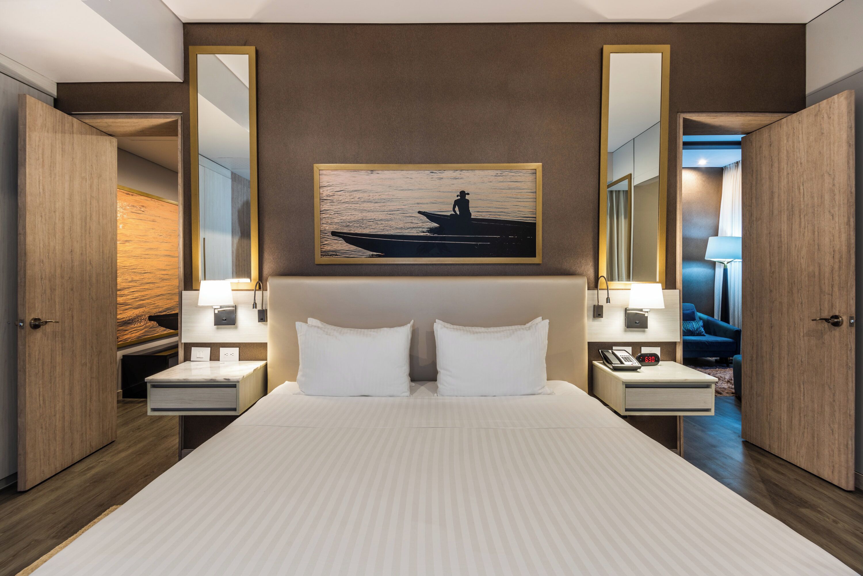 A hotel room with a large bed, two bedside tables with lamps, a nightstand with a phone, a painting above the bed, and a door leading to another room.