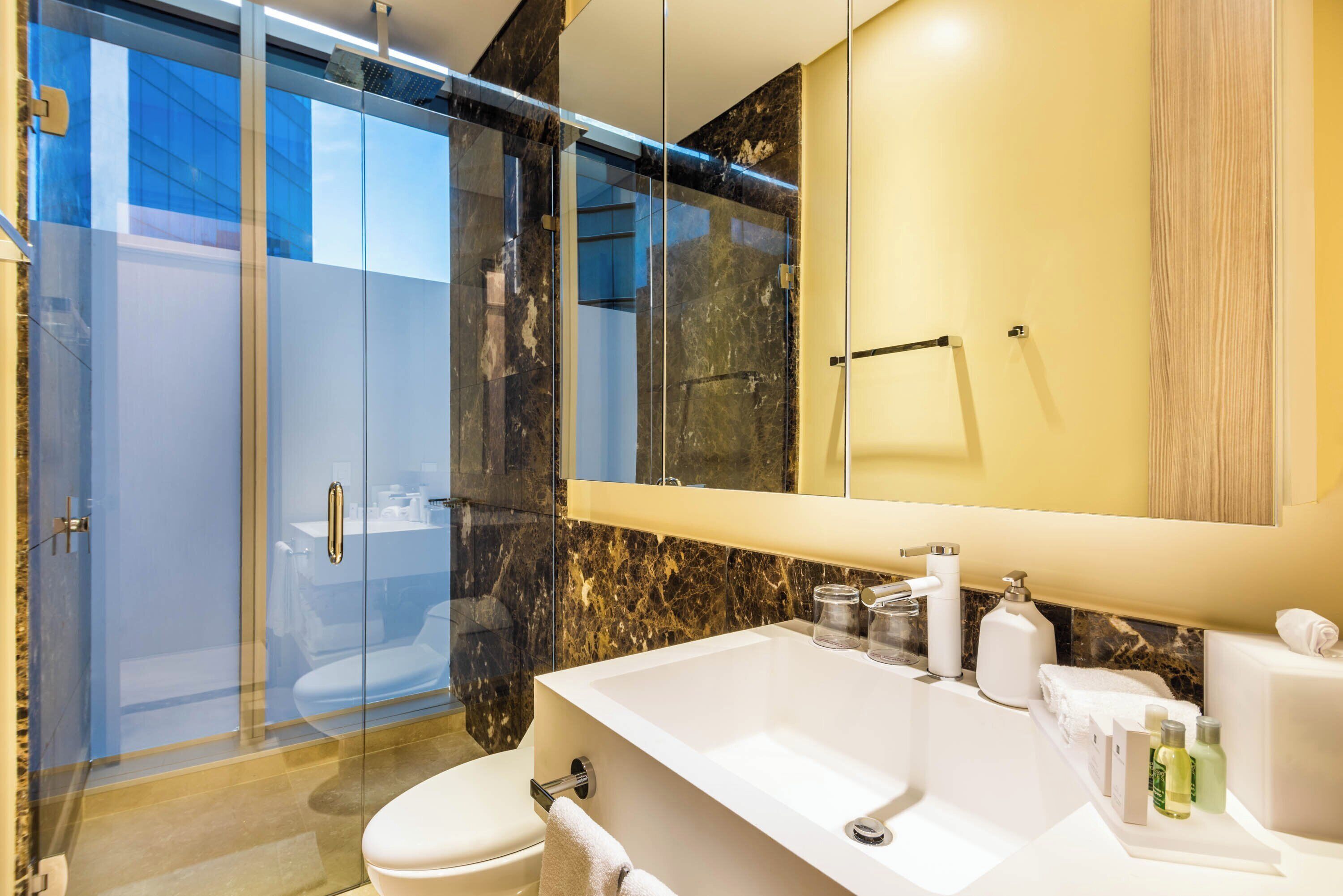 Un baño moderno con ducha envuelta en vidrio, un espejo grande y un lavamanos blanco con artículos de aseo.