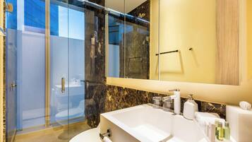 Un baño moderno con ducha envuelta en vidrio, un espejo grande y un lavamanos blanco con artículos de aseo.