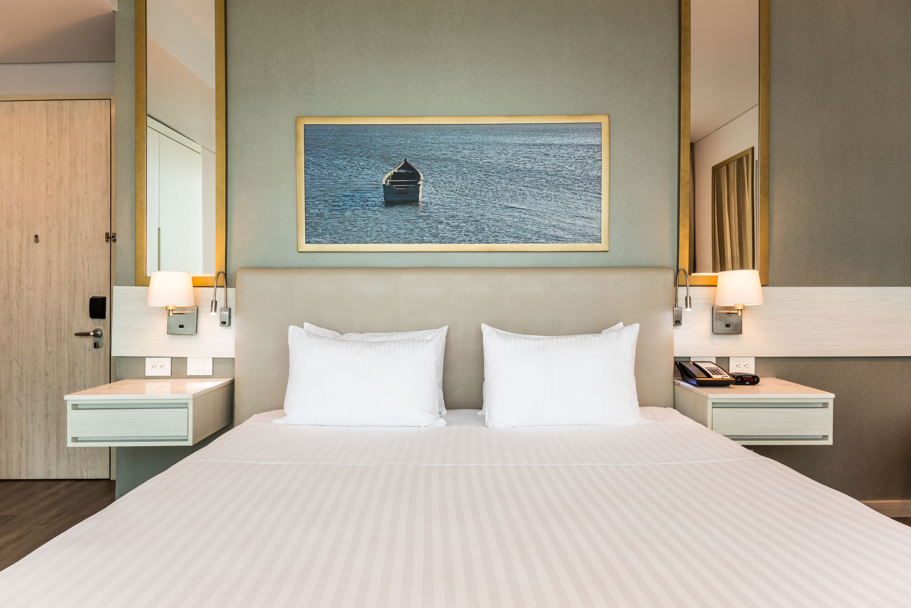 A hotel room with a large bed, two bedside tables with lamps, a wall-mounted painting of a boat on water, and a wooden door.