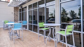 An outdoor seating area with tables and chairs, a stone wall, and large windows reflecting the outside. - Holiday Inn Express & Suites Siloam Springs by IHG (Siloam Springs)