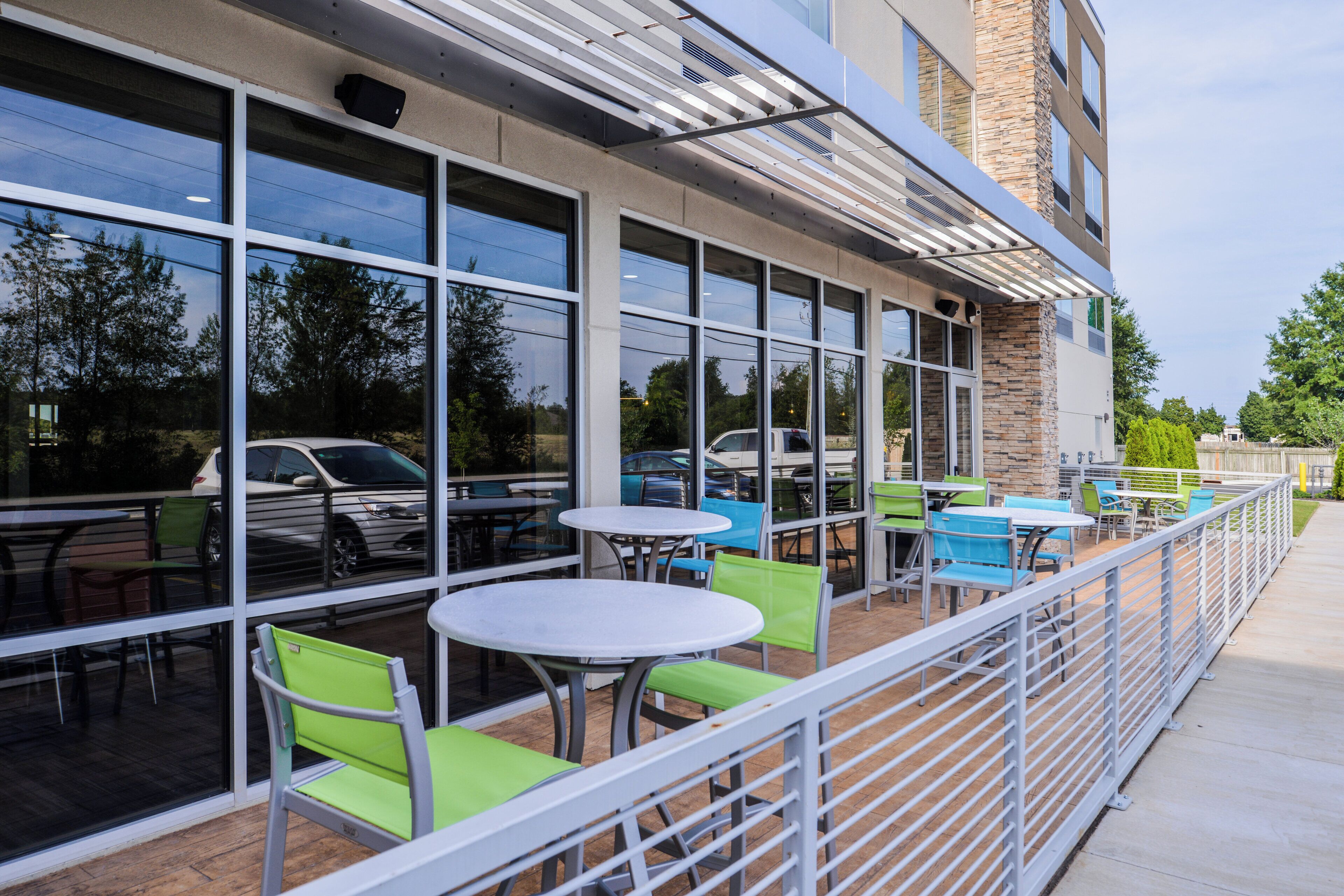 An outdoor seating area with tables and chairs, a stone building facade, and a view of parked cars and trees.