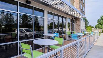 An outdoor seating area with tables and chairs, a stone building facade, and a view of parked cars and trees.