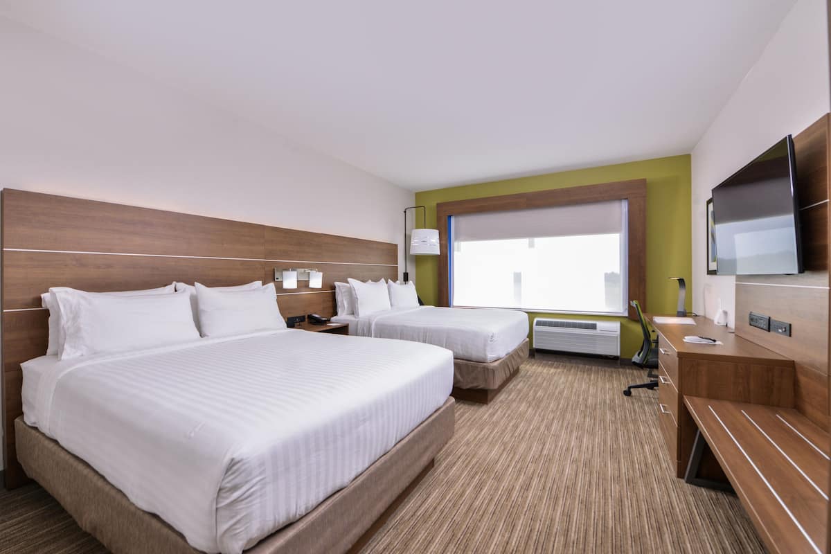 A hotel room with two beds, a TV, a desk, and a window with blinds.