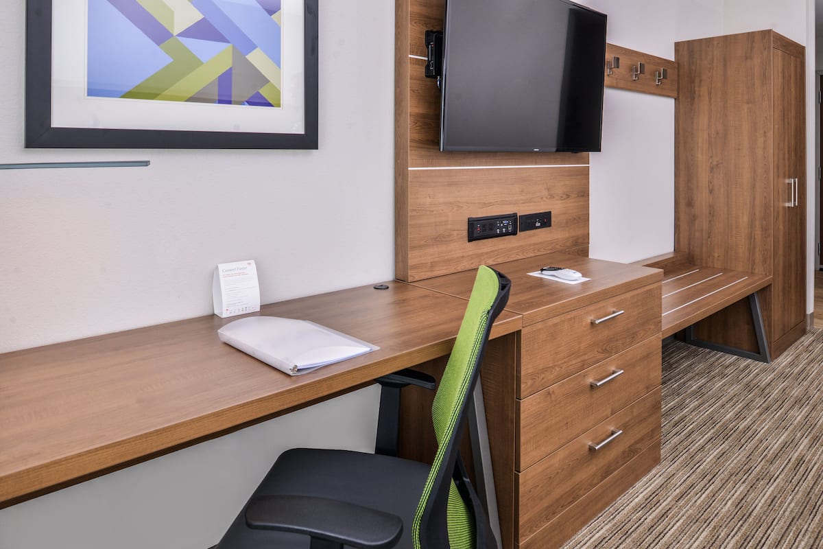 A hotel room with a wooden desk, a black office chair, a flat-screen TV mounted on the wall, and a wardrobe with a mirror.