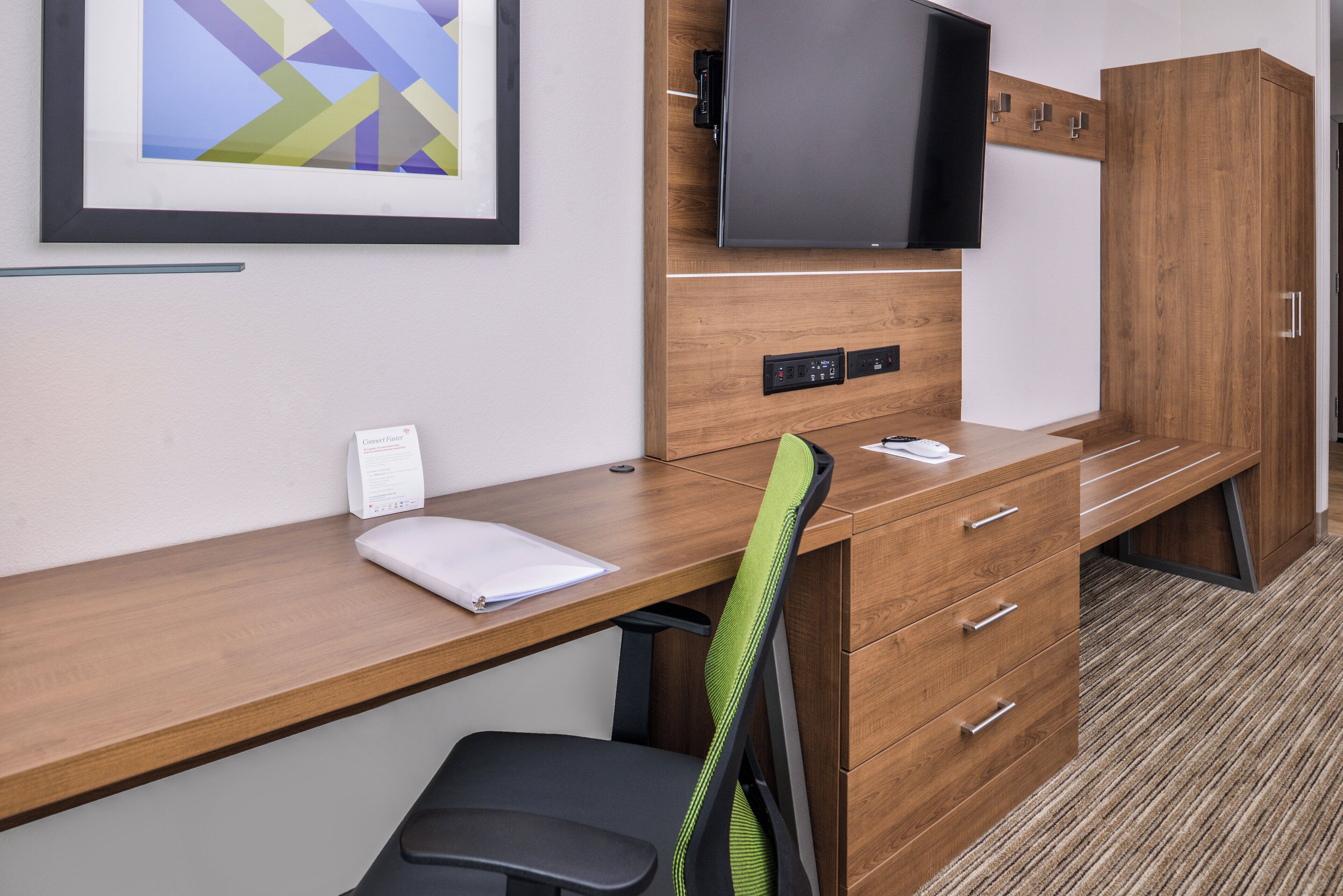Una habitación de hotel con un escritorio de madera, una silla, un televisor fijado a la pared y un cuadro enmarcado sobre el escritorio.