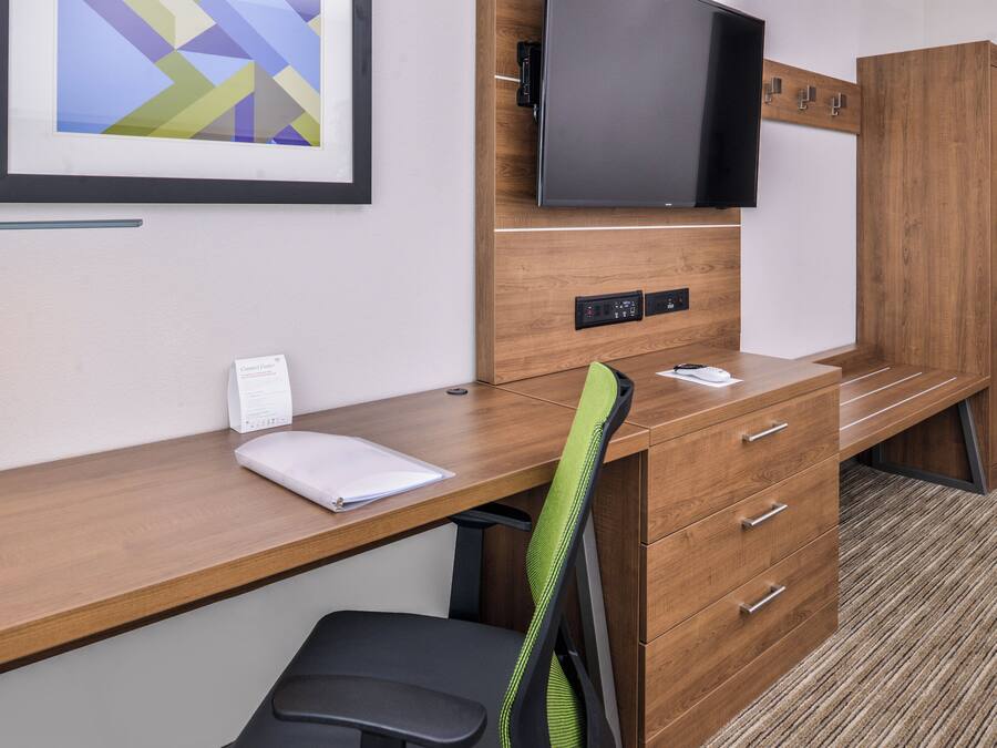 A hotel room with a wooden desk, a chair, a television mounted on the wall, and a framed picture above the desk.