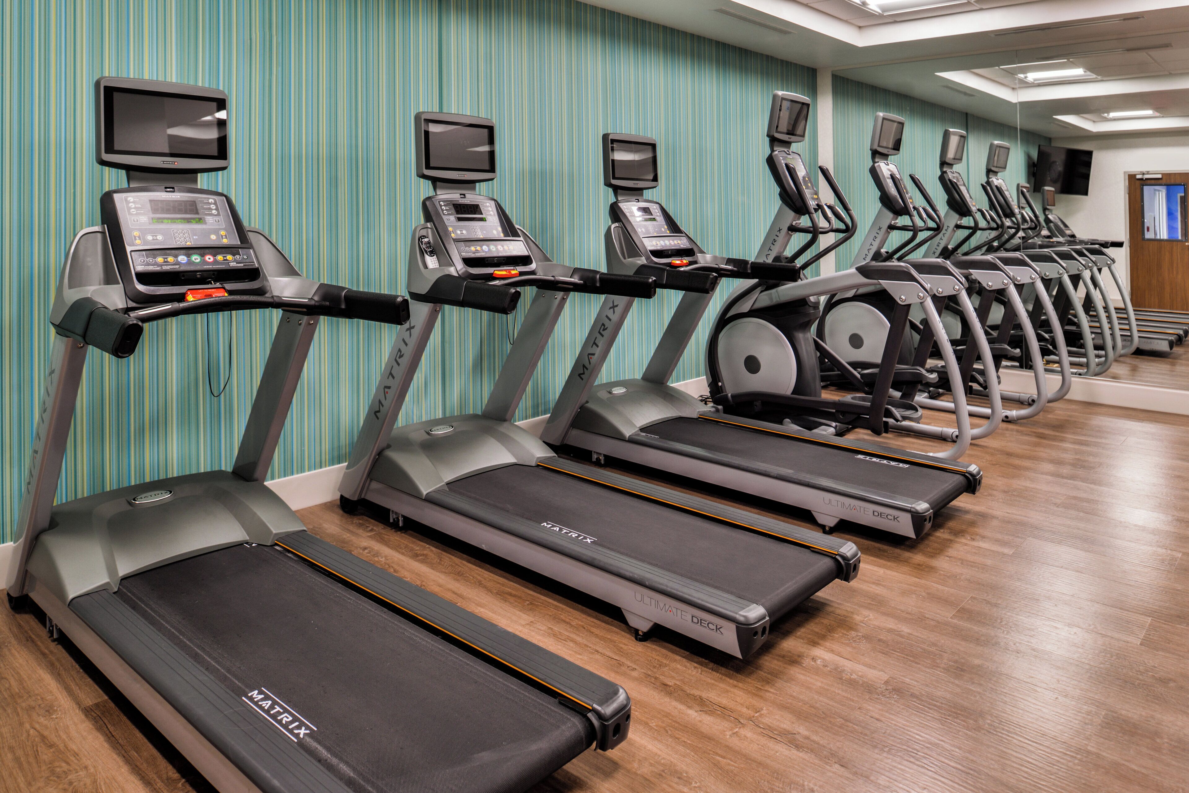 A row of treadmills with digital displays, arranged in a gym with striped wallpaper and wooden flooring.
