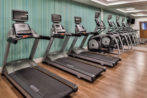 Una hilera de cintas de correr con pantallas digitales, dispuestas en un gimnasio con papel tapiz a rayas y suelo de madera.