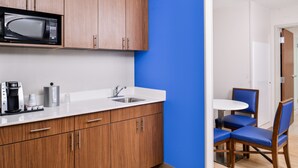 A modern kitchen with wooden cabinets, a microwave, a sink, and a coffee maker. There is a blue accent wall and a dining area with blue chairs. - Holiday Inn Express & Suites Siloam Springs by IHG (Siloam Springs)