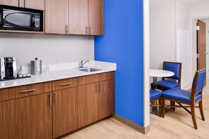 A modern kitchen with wooden cabinets, a microwave, a sink, and a coffee maker. There is a blue accent wall and a dining area with blue chairs. - Holiday Inn Express & Suites Siloam Springs by IHG (Siloam Springs)