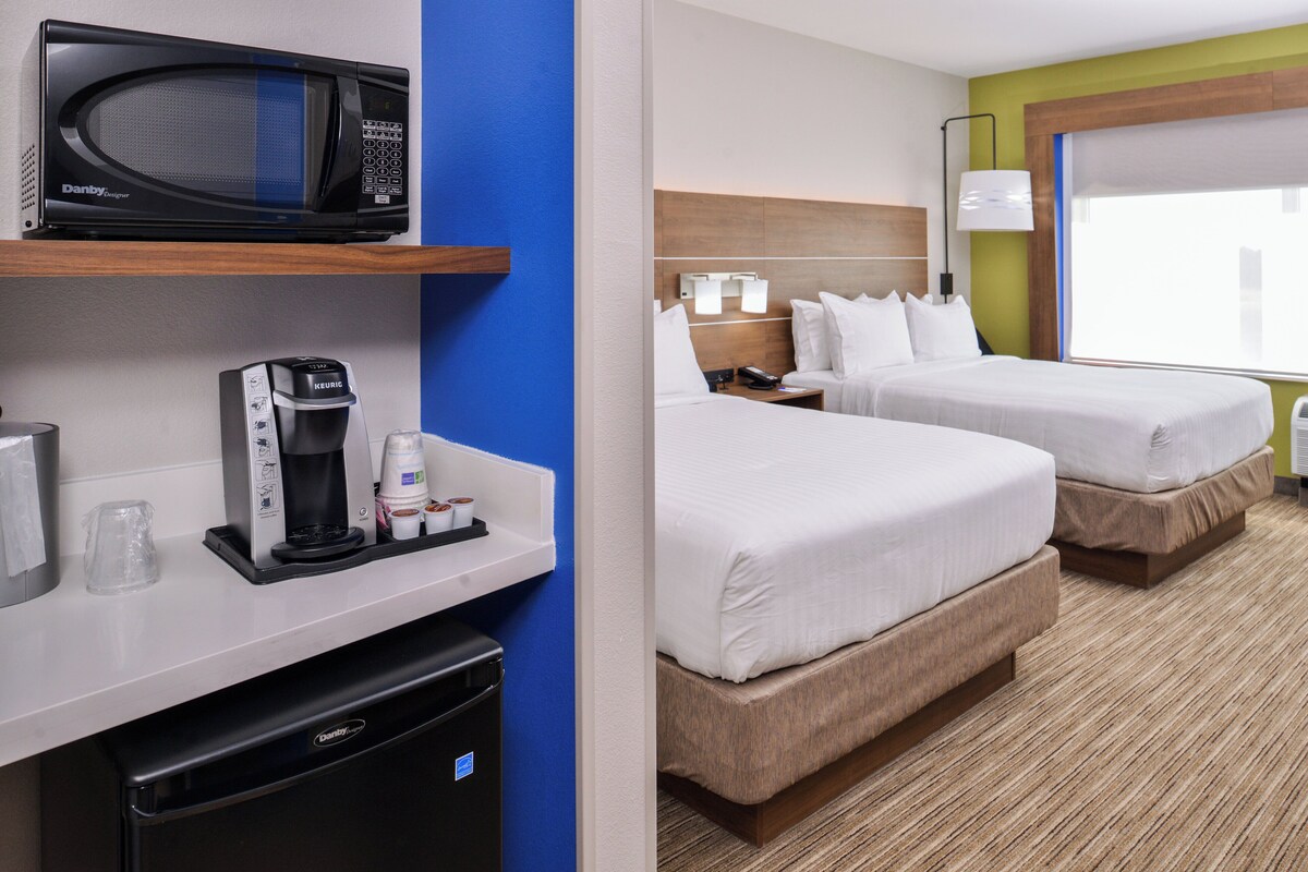A hotel room with a microwave, coffee maker, and a compact refrigerator. There are two beds with white linens and a large window providing natural light.