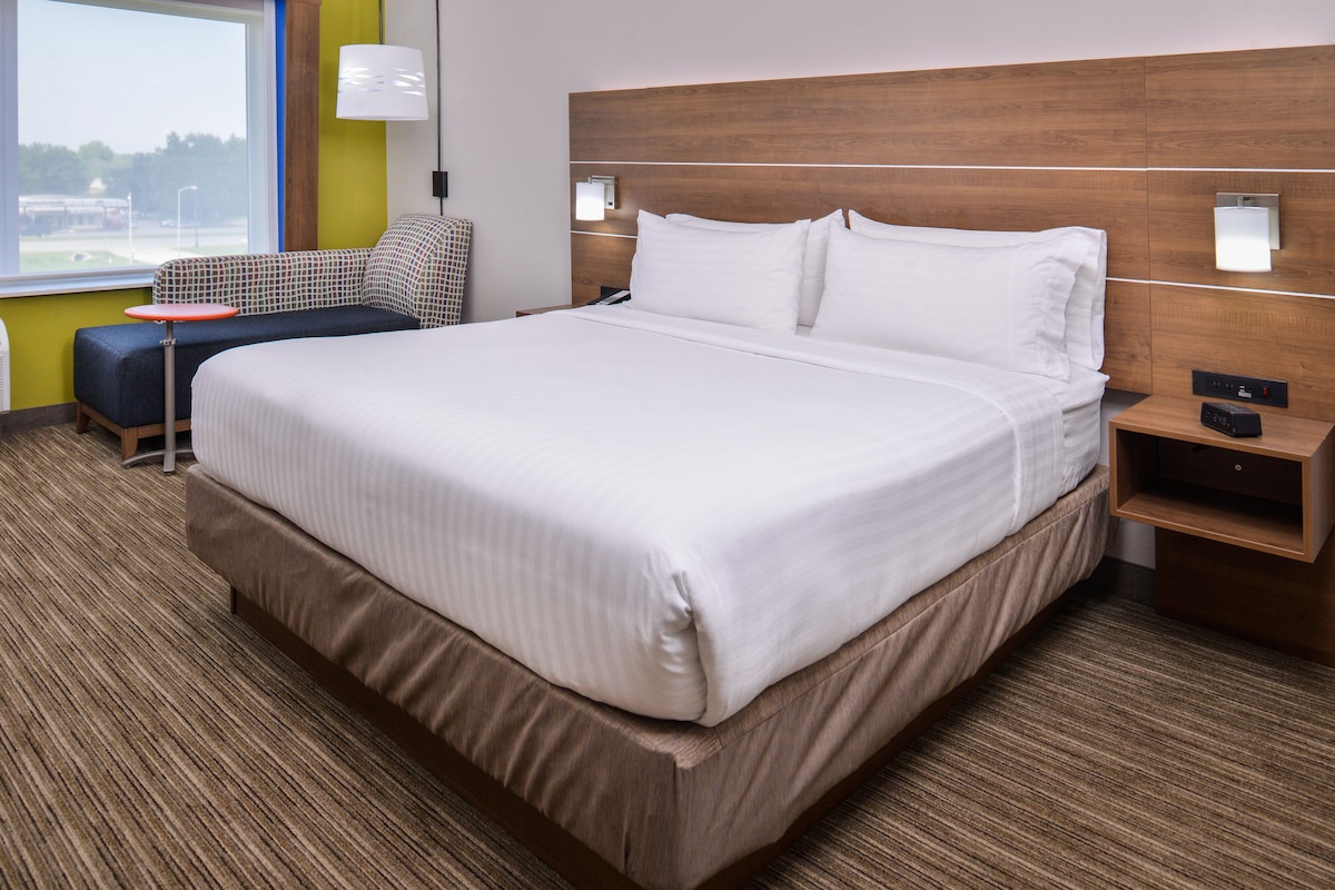 Habitación de hotel con una cama grande, una mesita pequeña, una silla y una mesita de noche con lámpara.