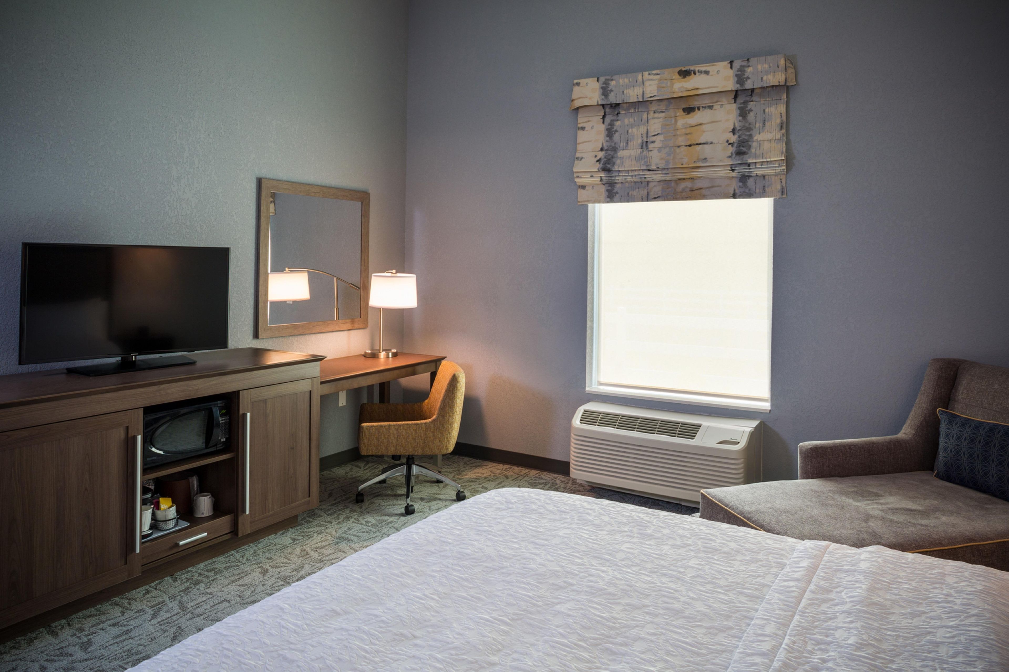 Une chambre d’hôtel comprenant un lit, un bureau avec une chaise, une télévision, un miroir et une fenêtre avec des stores.