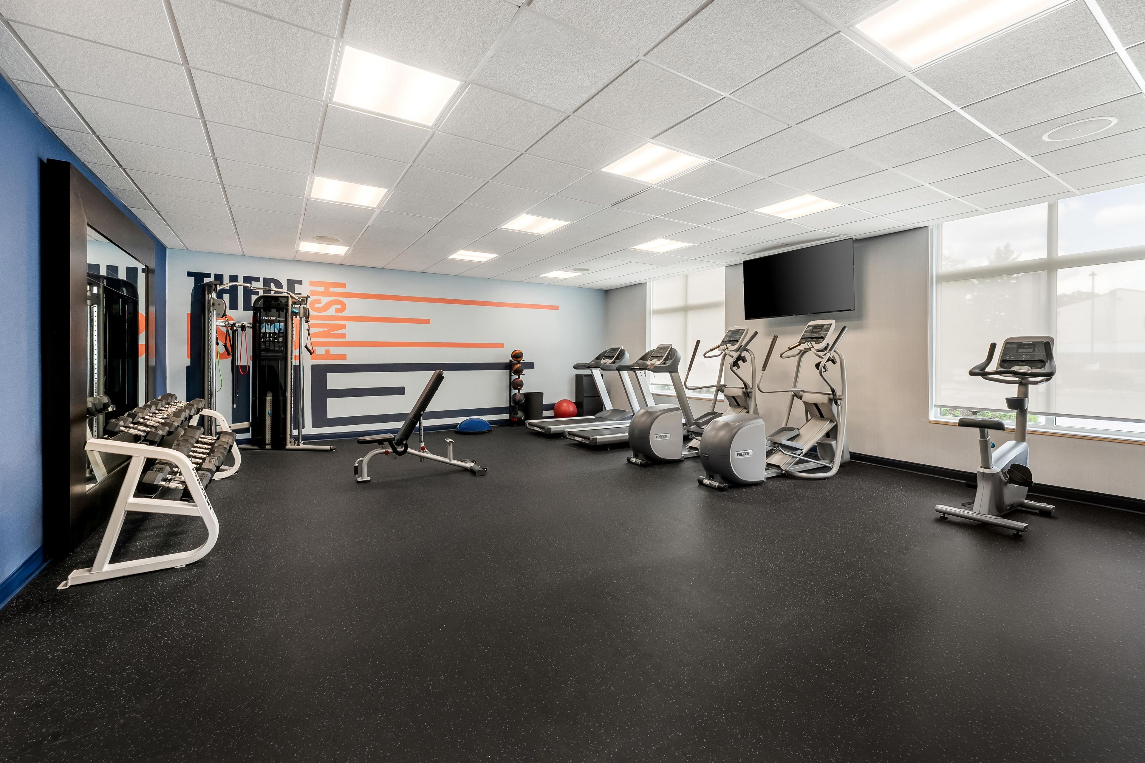 A gym with various exercise equipment, a wall with motivational text, and large windows.