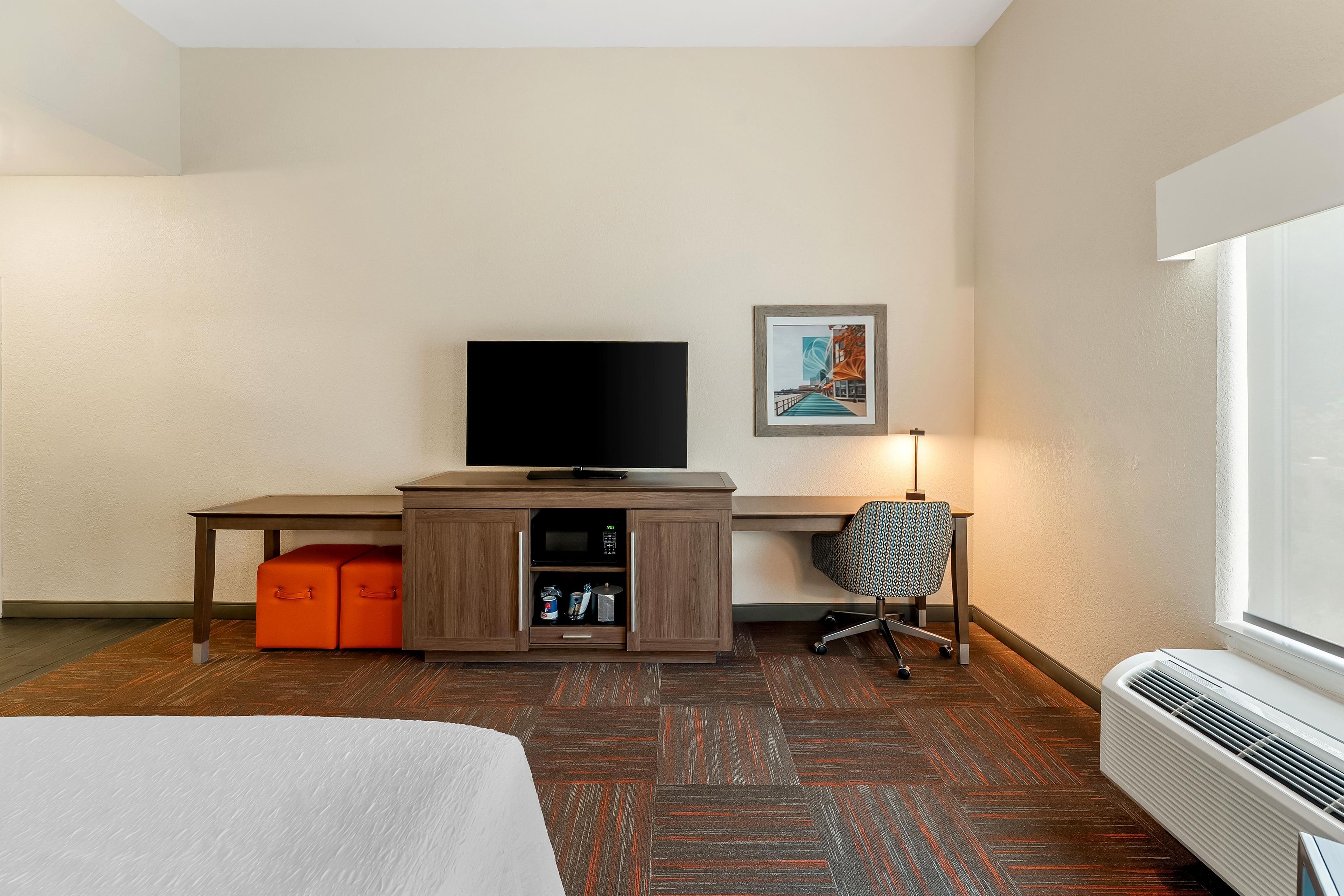 Habitación de hotel con cama, escritorio con televisor, silla, mueble pequeño y ventana.