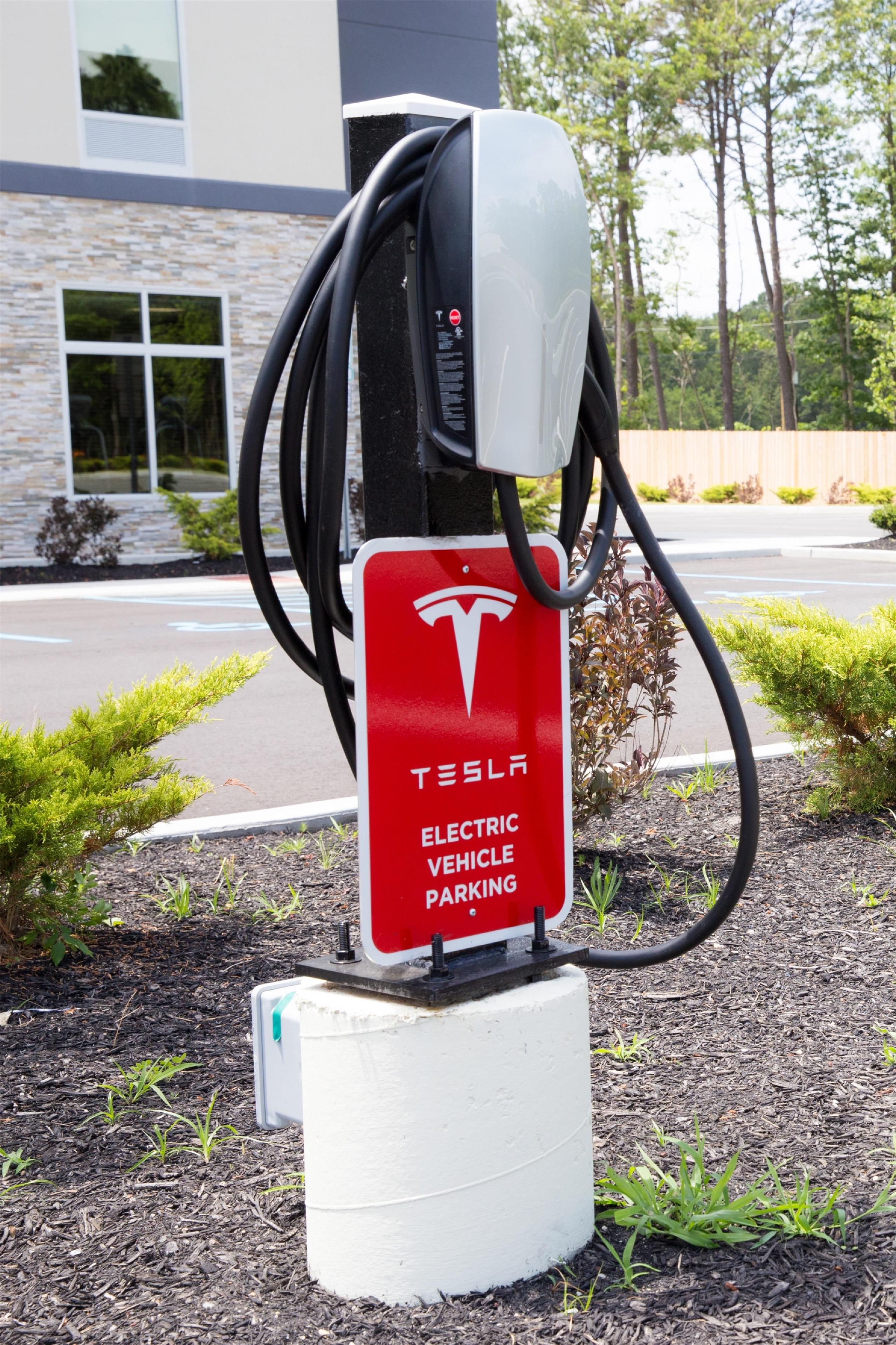 A Tesla electric vehicle charging station with a red sign, located outdoors with a building and trees in the background.