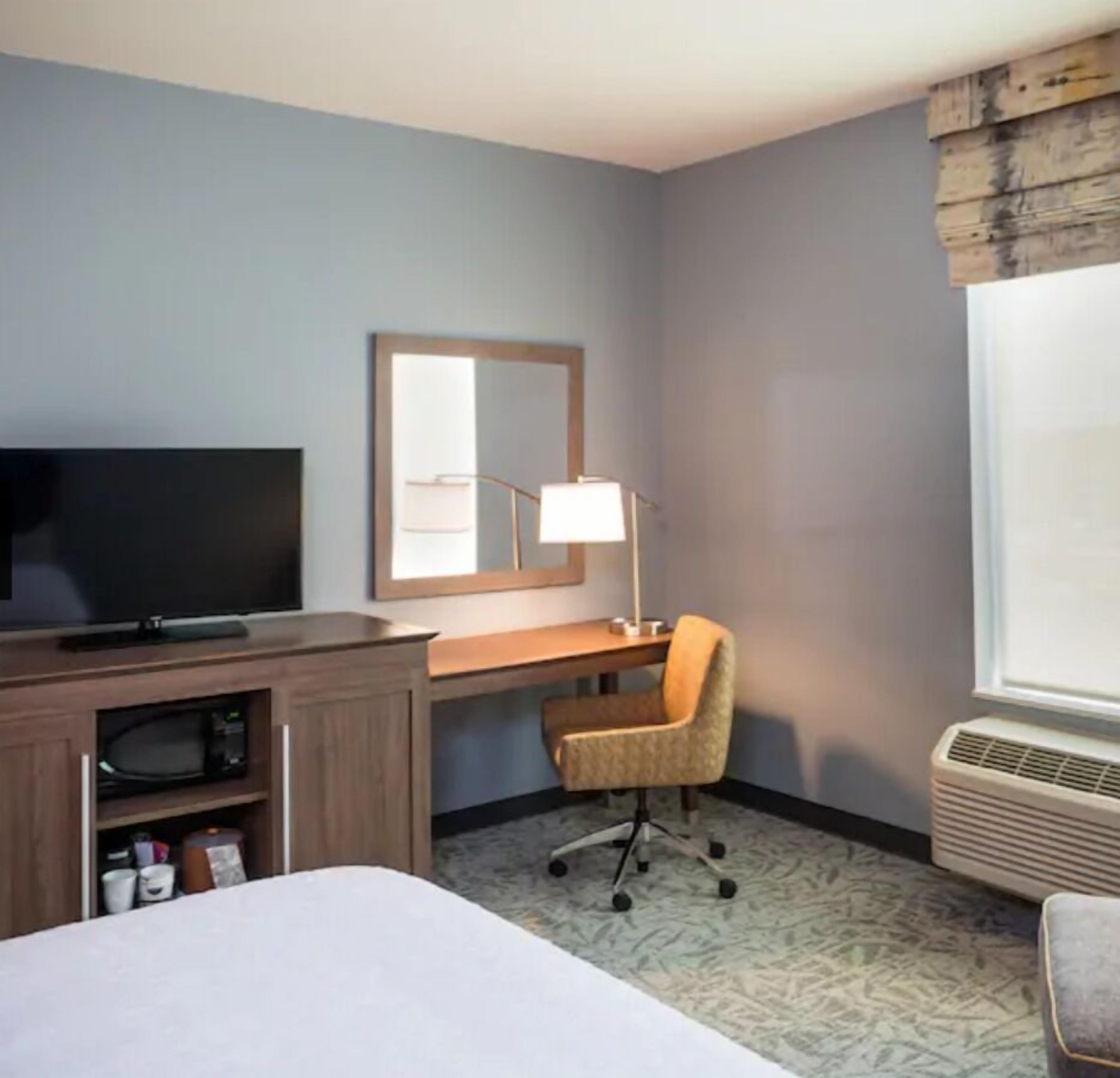 Ein Hotelzimmer mit Bett, Schreibtisch mit Stuhl, Fernseher, Spiegel und Fenster mit Vorhängen.