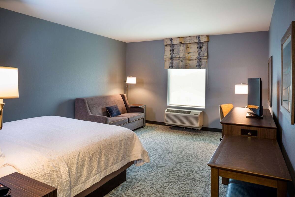 A hotel room with a bed, a sofa, a desk, a TV, a window, and an air conditioning unit.