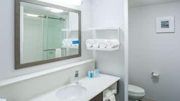 A modern bathroom with a large mirror, white sink, and a toilet. There are shelves with towels and toiletries.
