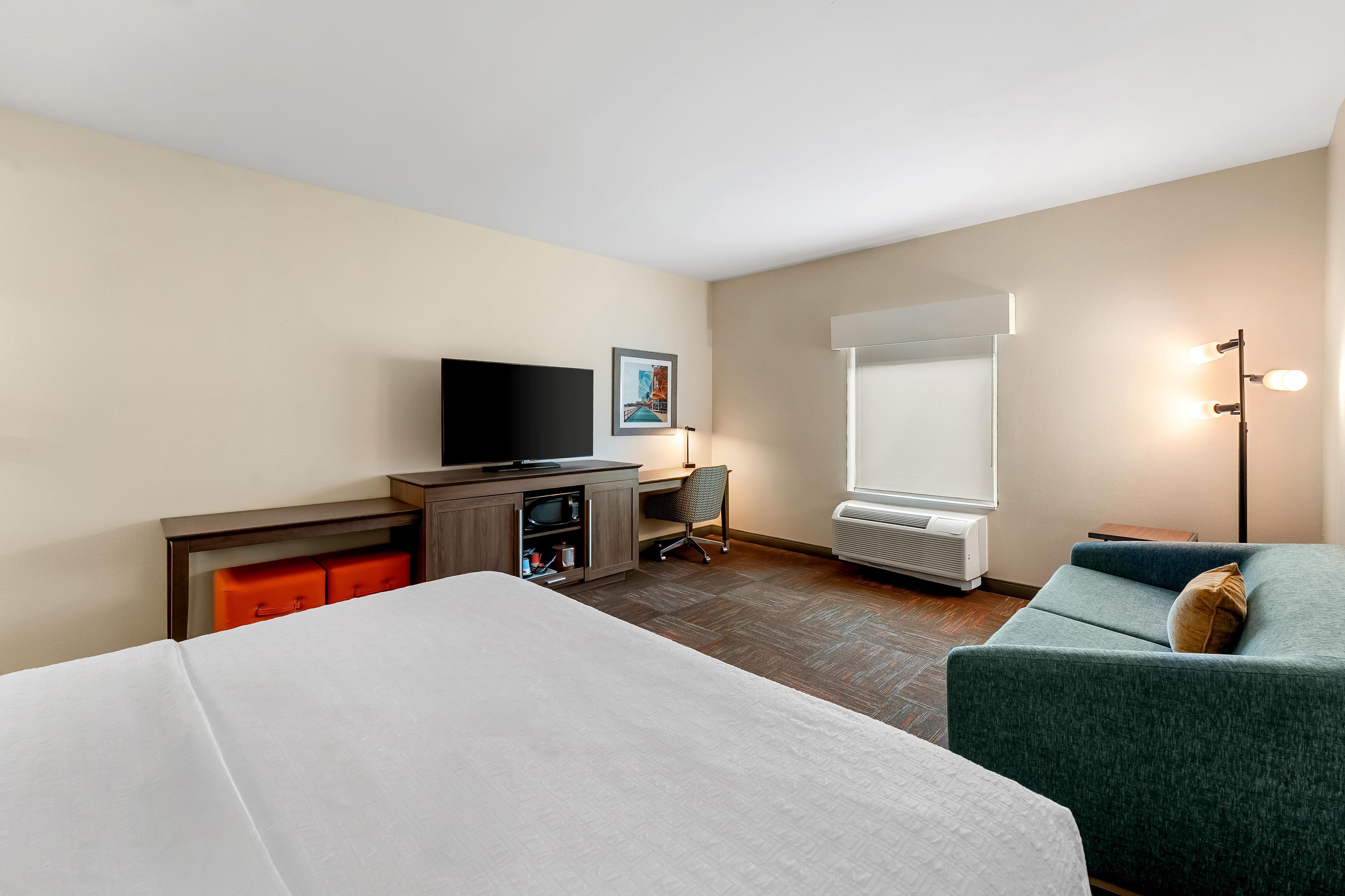 A hotel room with a bed, a desk with a television, a chair, a sofa, and a window with blinds.