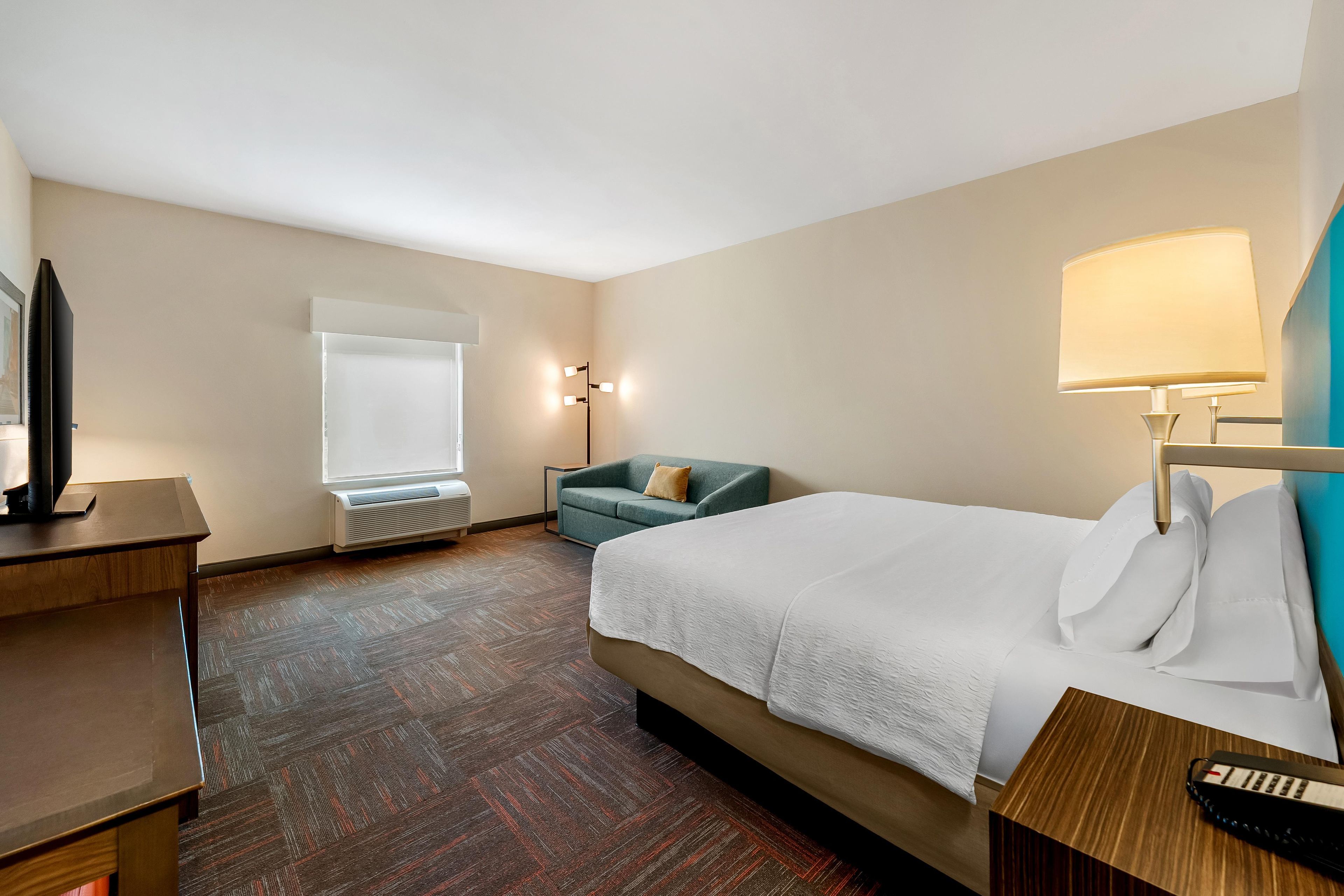 A hotel room with a bed, a desk, a chair, a standing lamp, and a window.
