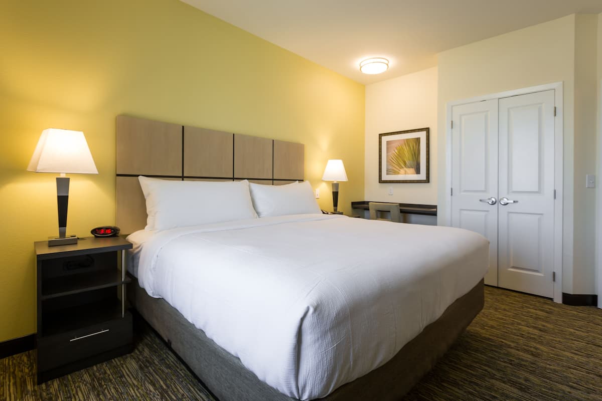 Una habitación de hotel con una cama grande, dos mesitas de noche con lámparas, una mesita auxiliar y un cuadro enmarcado en la pared, además de una puerta.