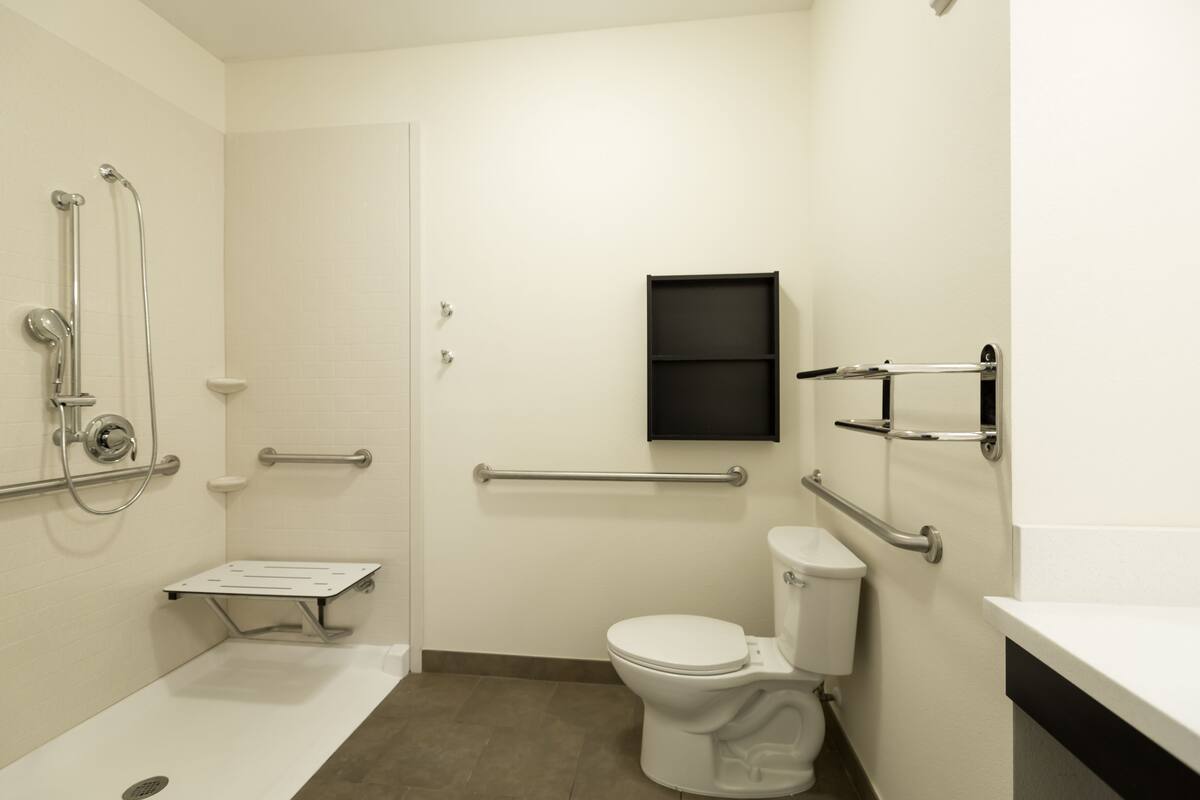 Un baño moderno con inodoro blanco, barras de apoyo, ducha con cabezal de ducha manual y un estante pequeño.