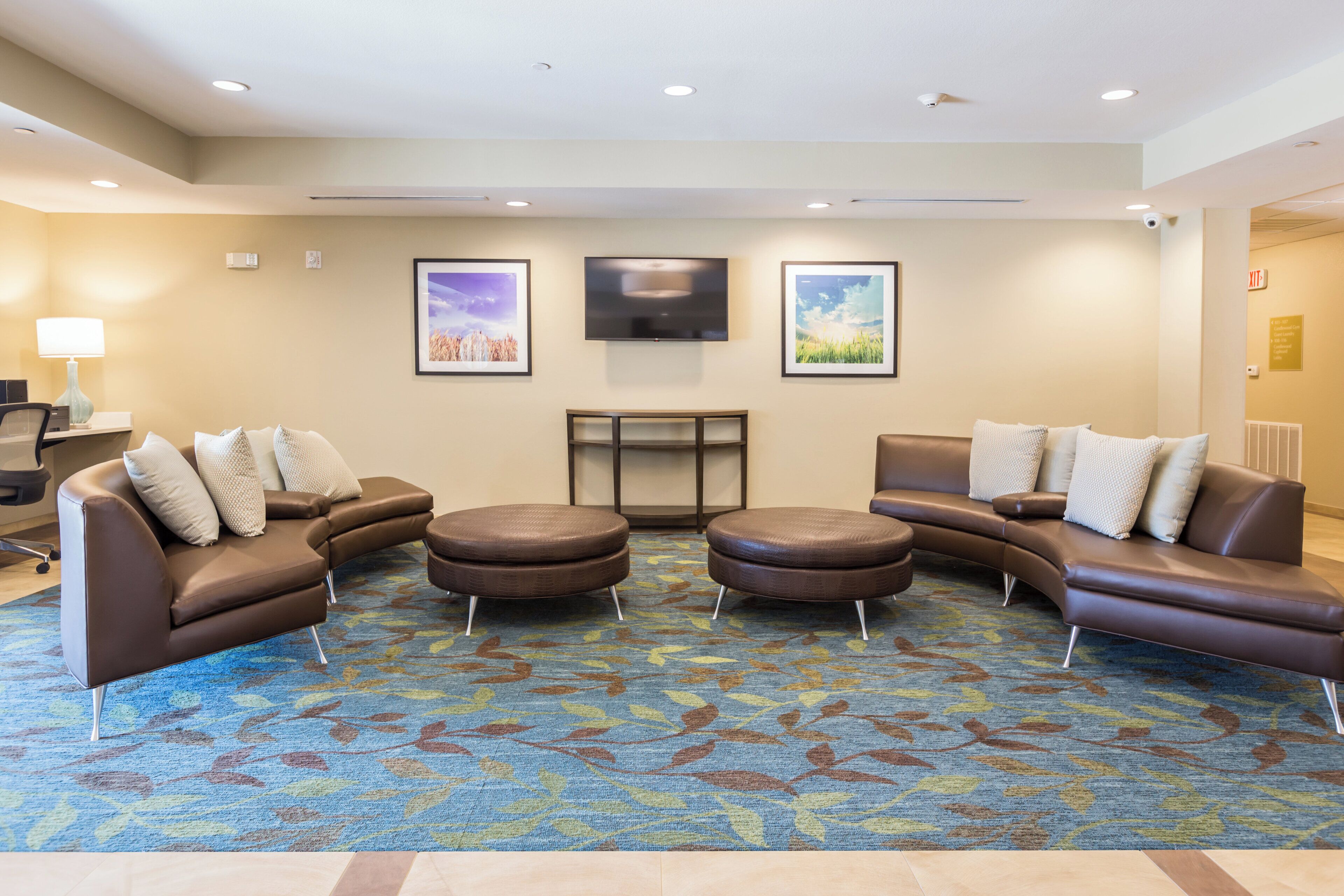 A hotel lobby with a seating area, a flat-screen TV mounted on the wall, and a carpeted floor.
