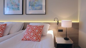Een netjes opgemaakt bed met witte en oranje kussens, een nachtkastje met een lamp, en twee kunstwerken in lijsten aan de muur.