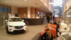 Een moderne hotellobby met een witte auto op de voorgrond, een receptiebalie en zitgedeeltes met oranje en witte stoelen.