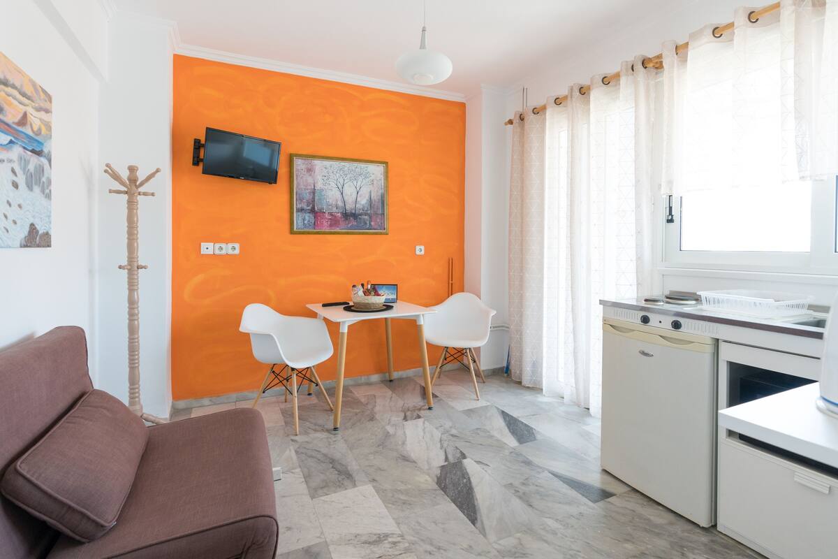 A compact living space with a sofa, a dining table with chairs, a television mounted on an orange wall, and a kitchen area with a dishwasher and stove.