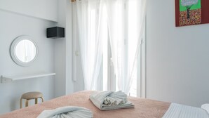A neatly made bed with folded towels, a round window, a wall-mounted air conditioner, and sheer curtains. - Poseidon Apartments (Kefalonia)
